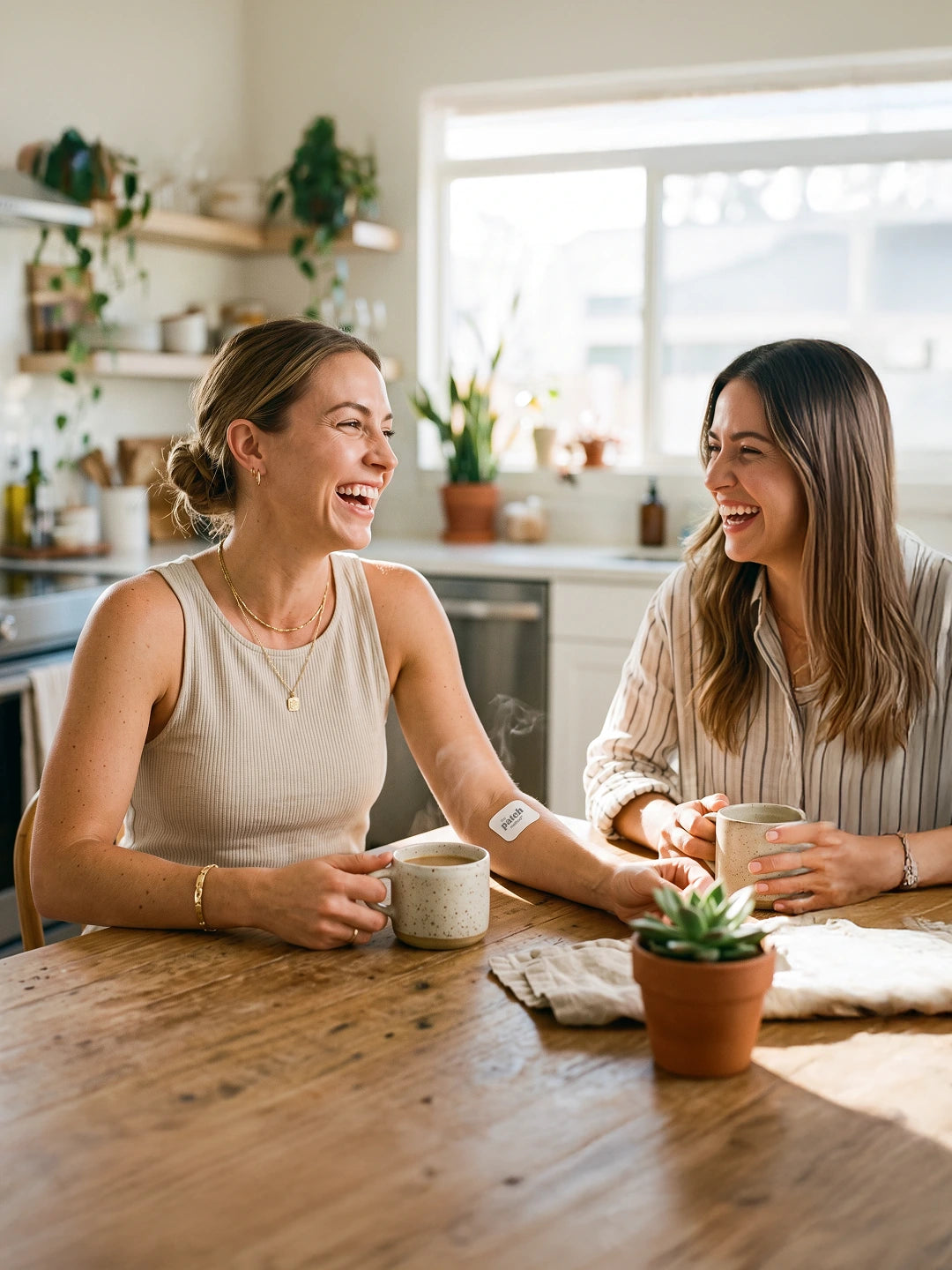 Two women laughing together at a kitchen table over coffee, one with a Patch Method patch visible on her forearm
